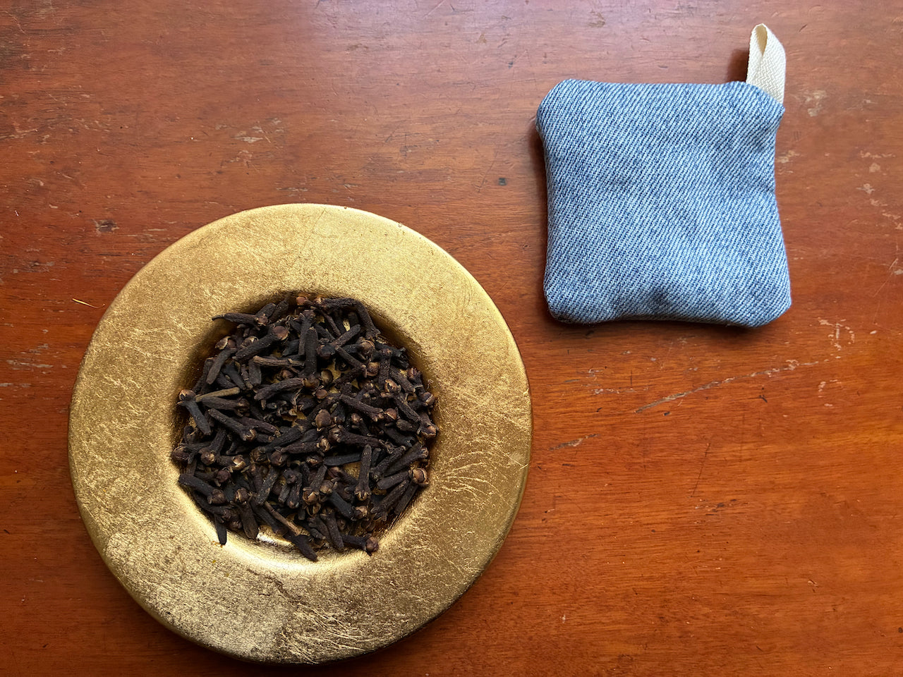 One Blue Denim Clove Sachet and a saucer of Whole Cloves on a wooden surface