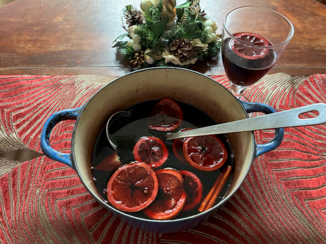 Mulled wine in a pan with sliced oranges and cinnamon sticks on a table with a glass of mulled wine.
