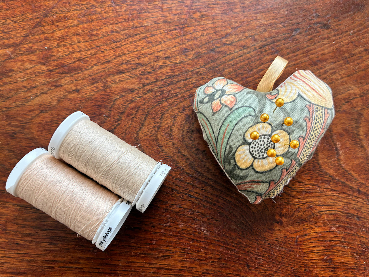 Small William Morris Pin Cushion with two reels of Cream Thread on a wooden surface