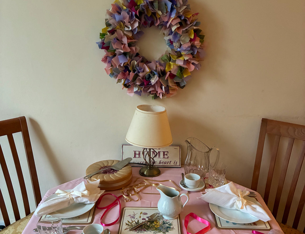 Dining table set with a pink tablecloth, plates, and cutlery, with a decorative wreath on the wall.