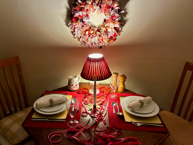 Romantic dining table set for two with a colourful circular Valentine Wreath on the wall behind.