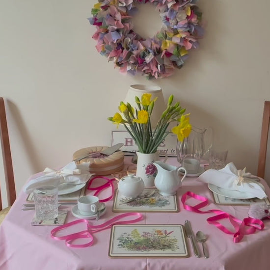 Circular wreath in pinks, yellows, greens and lilacs beside a dining table with a vase of daffodils  and spring tableware