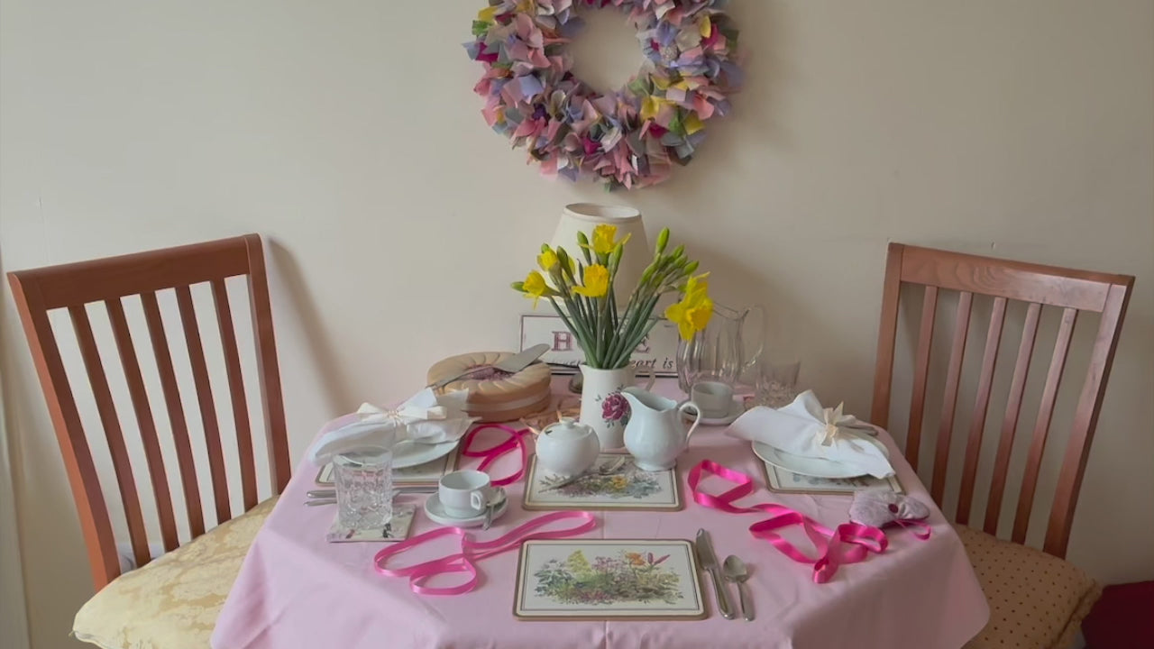 Circular wreath in pinks, yellows, greens and lilacs beside a dining table with a vase of daffodils  and spring tableware