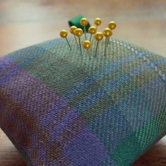 A Tartan Pincushion with Gold pins in it on a wooden surface