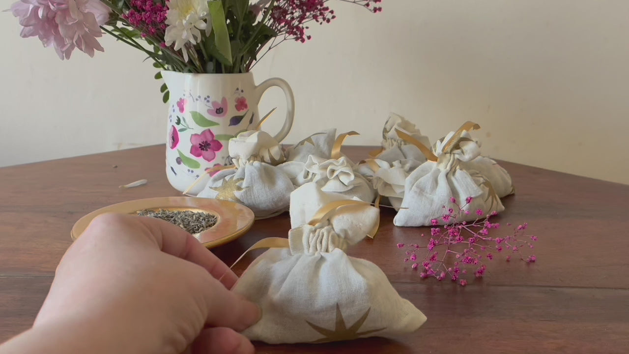 One Cream and Gold Starry Lavender Sachet by a saucer of Dried Lavender and a vase of Pink flowers